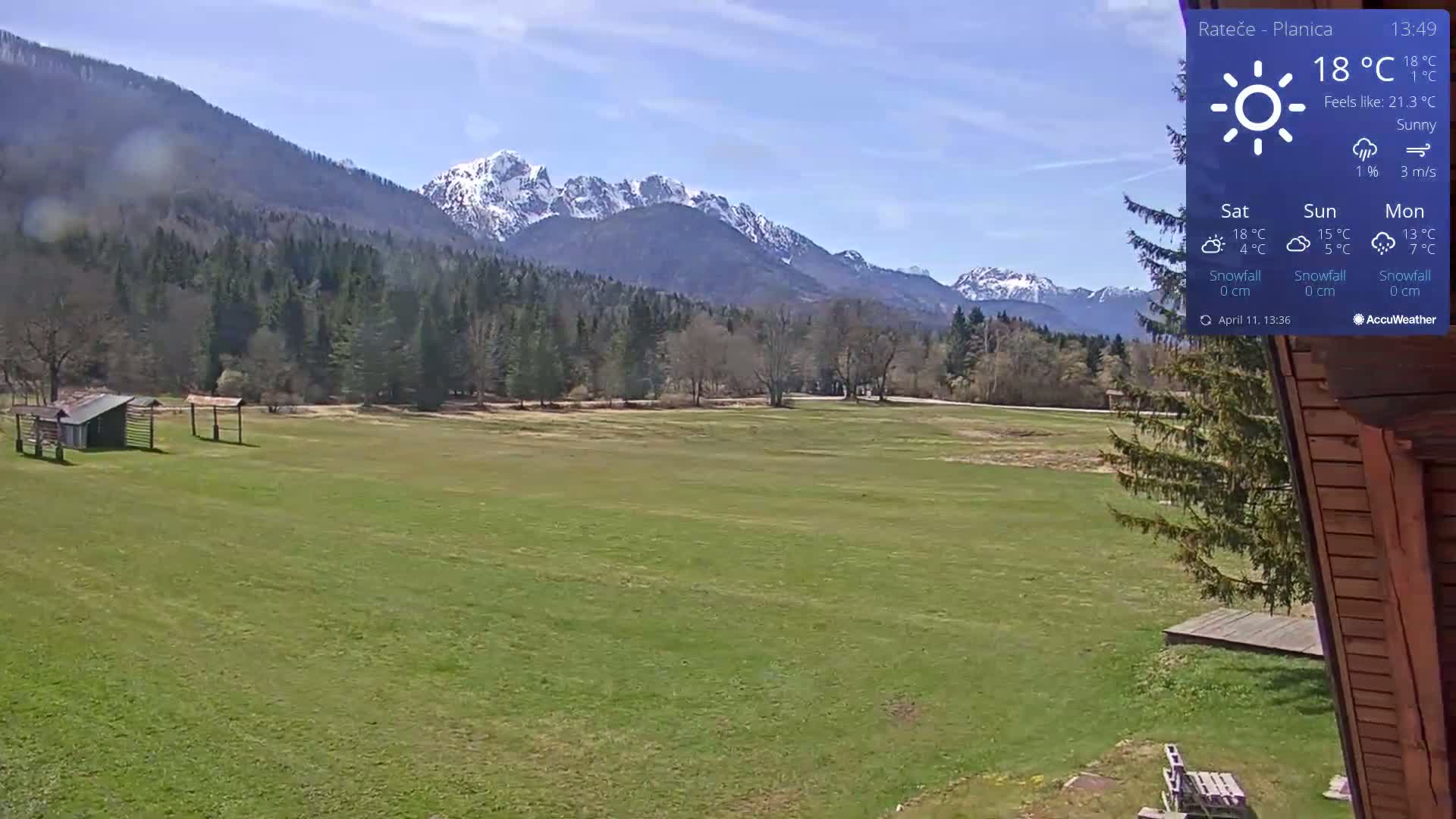 A sunny view of a grassy field with a few small structures, backed by a forest and snow-capped mountains.