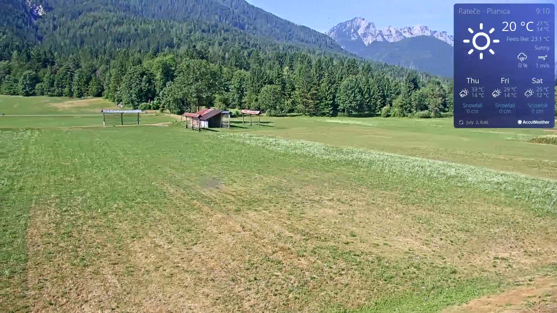 A sunny, expansive green field with two small sheds sits before a backdrop of a lush green forest and distant mountains.