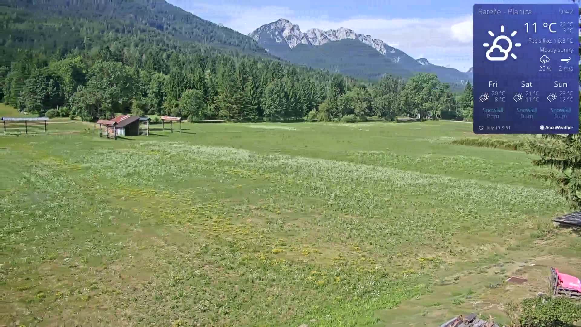 A mostly sunny, expansive green field dotted with small structures sits before a backdrop of dark green forested hills and distant snow-capped mountains.