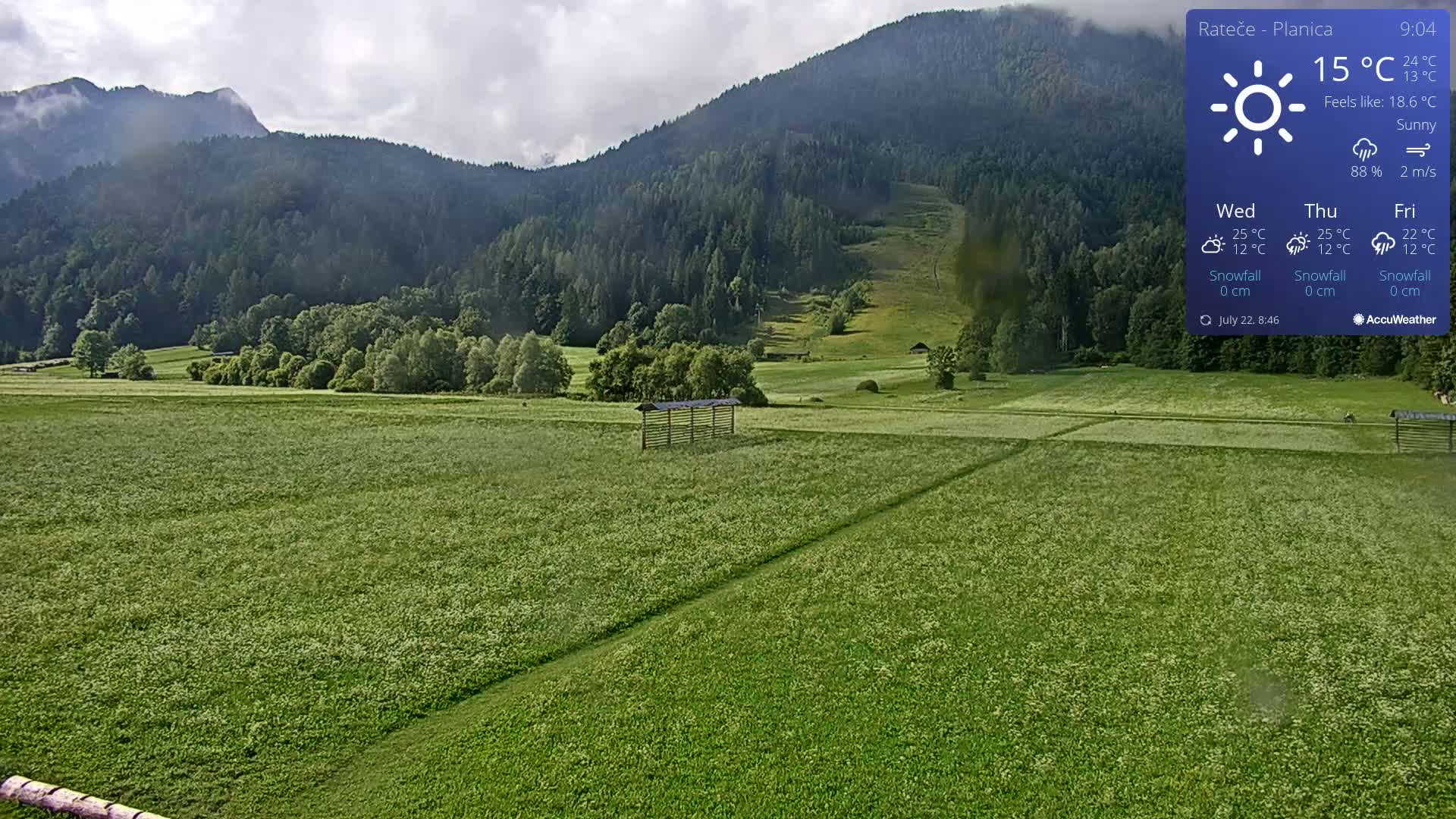 A sunny, green field stretches towards a mountainous backdrop under a partly cloudy sky.