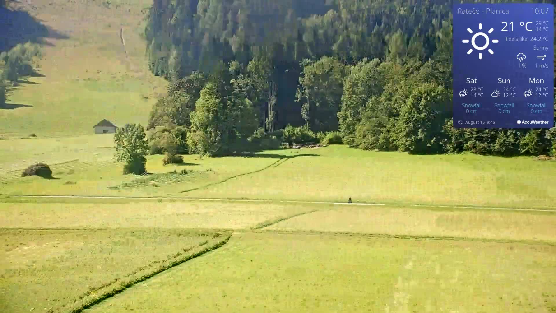 A sunny, high-angle view shows a vast, green field with a small building, trees, and a person in the distance, under a clear sky.