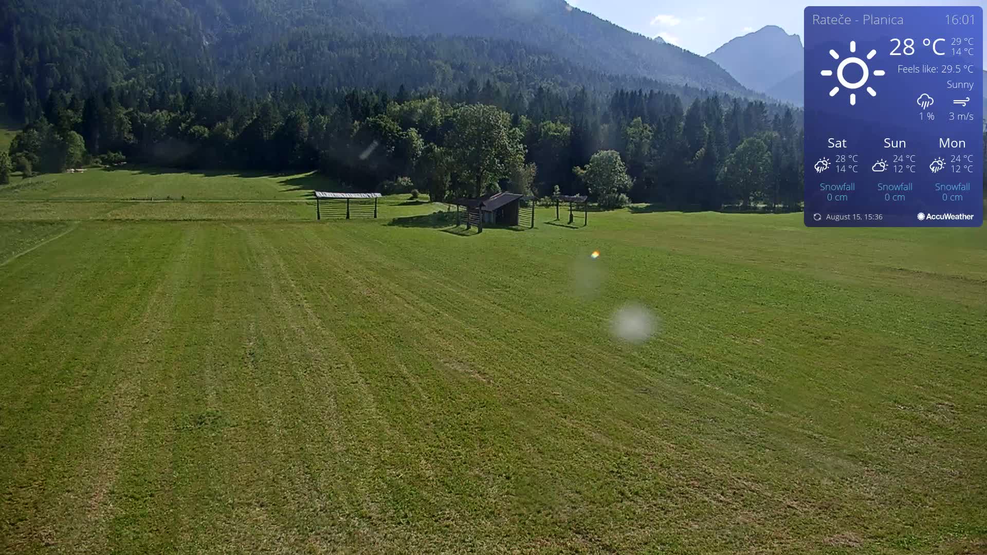 A sunny, high-angle view shows a vast, green field with a small building, trees, and a person in the distance, under a clear sky.