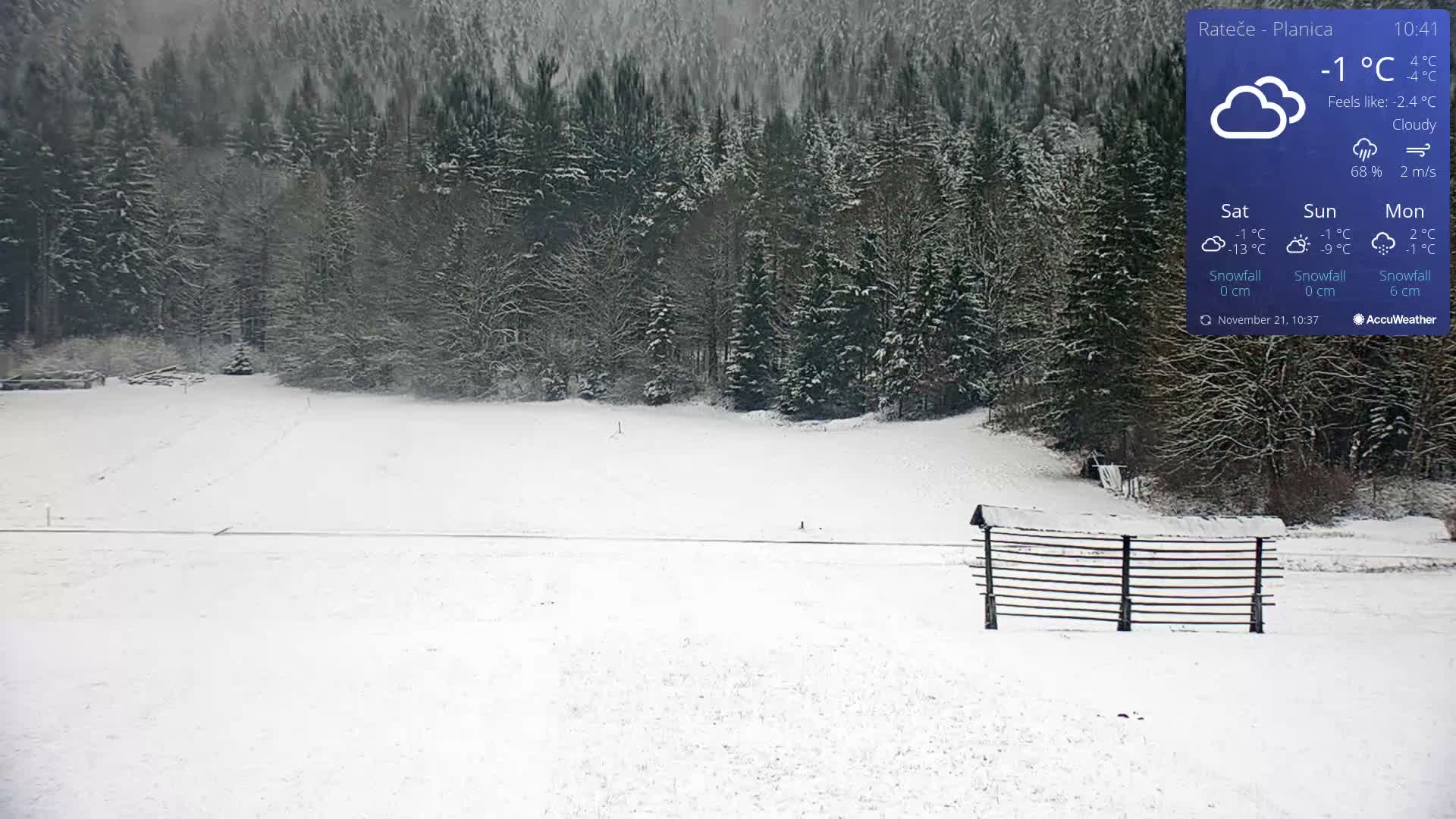 A wide, snow-covered field stretches towards a dense, snow-laden forest, with a traditional wooden hay rack standing prominently on the right, all under cloudy skies.
