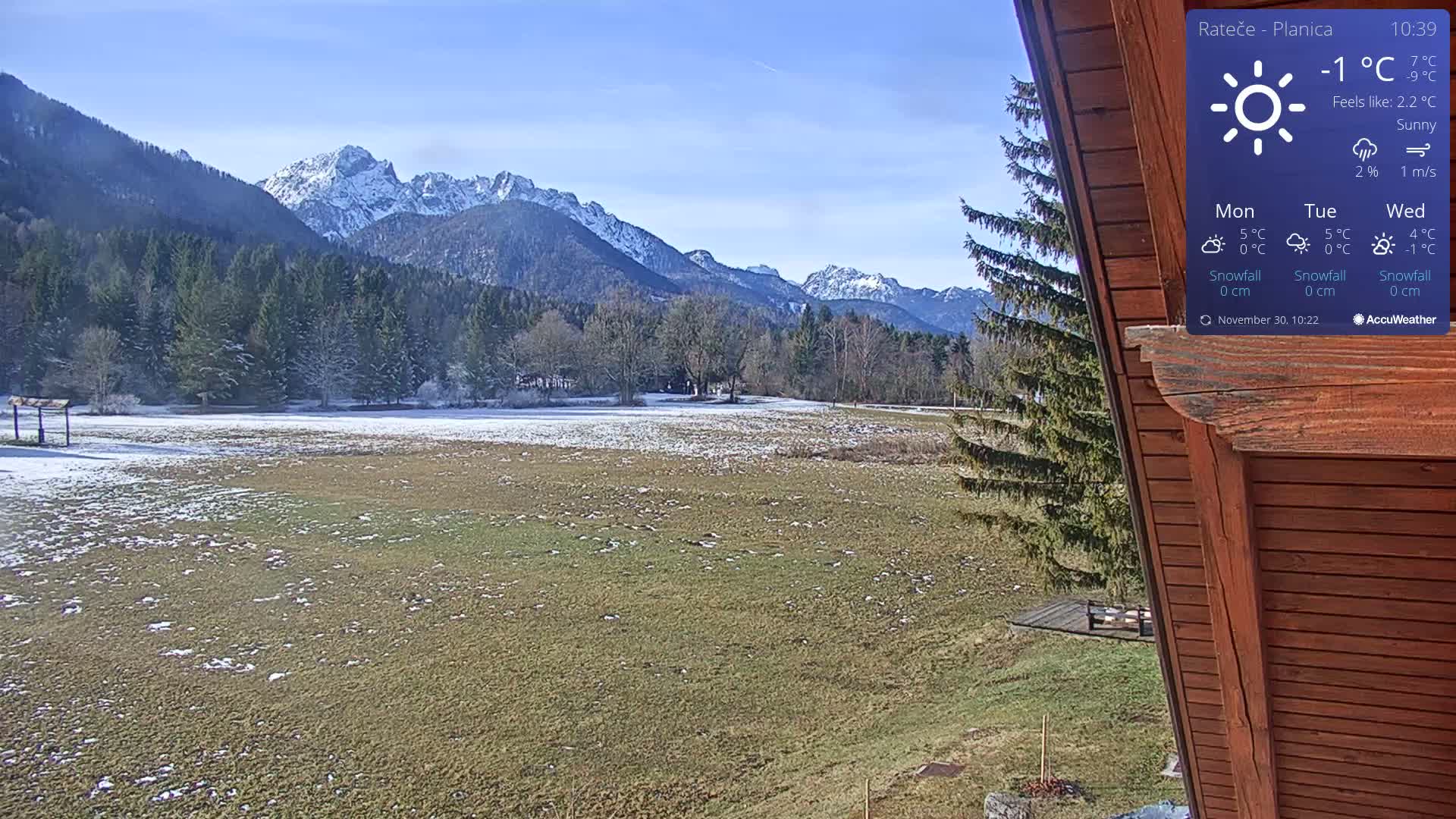 A sunny winter landscape showcases snow-capped mountains and a forest in the background, a partially snow-dusted green field in the middle ground, and a wooden building framing the scene on the right.