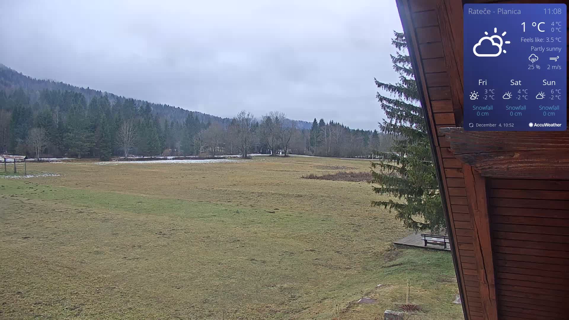 A wide, frosty field with patches of snow leads to a dense evergreen forest covering a misty hillside under a cloudy winter sky, viewed from the eaves of a wooden structure.
