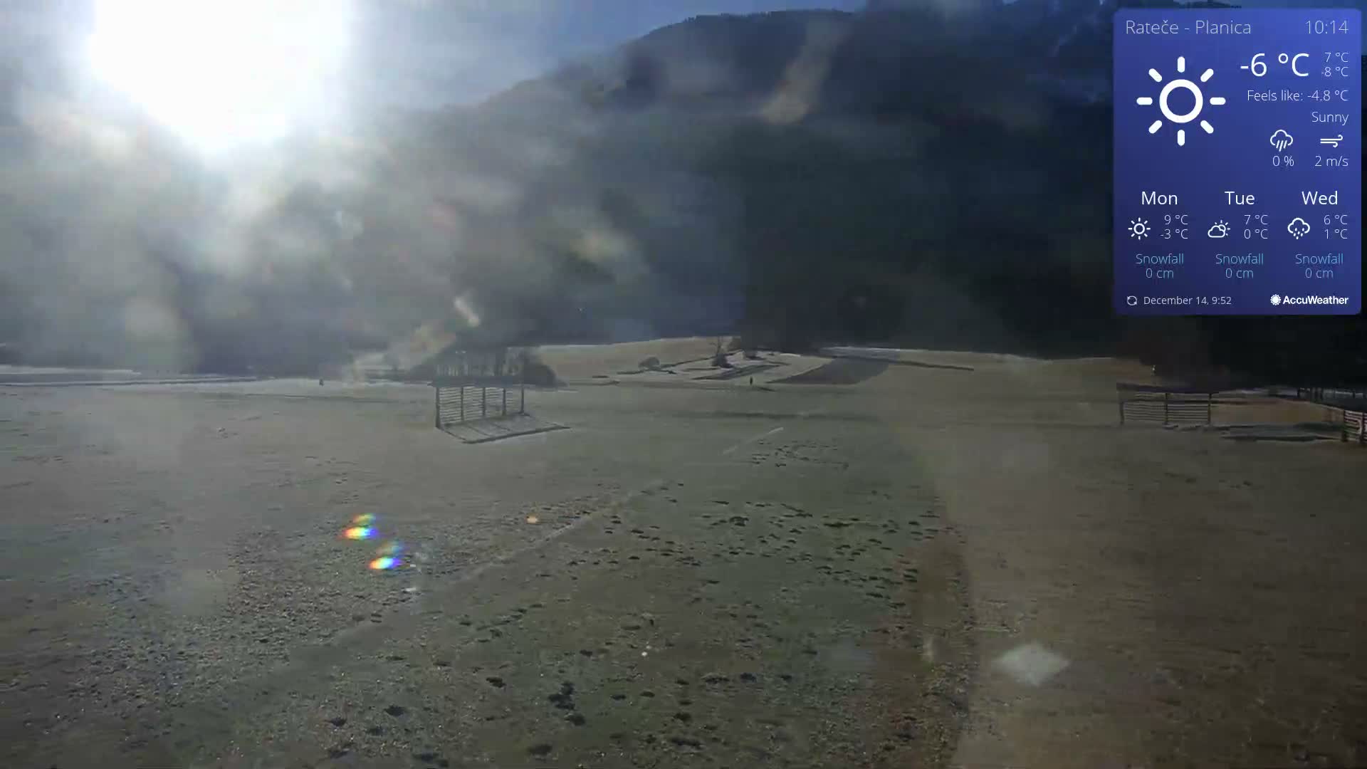 A wide, frosty field with patches of snow leads to a dense evergreen forest covering a misty hillside under a cloudy winter sky, viewed from the eaves of a wooden structure.