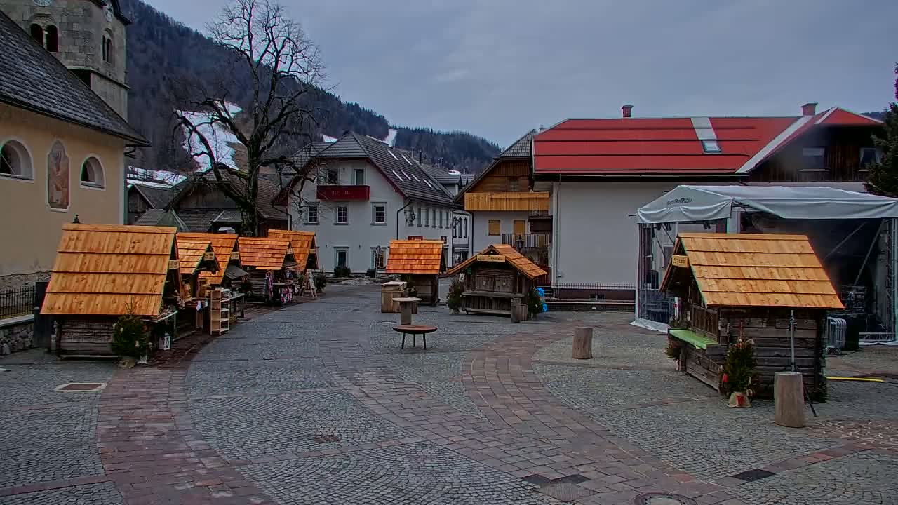 A cobblestone town square, under an overcast sky, features several small wooden market stalls with peaked roofs and a larger building with a red roof in the background; snow-covered mountains are visible in the distance.