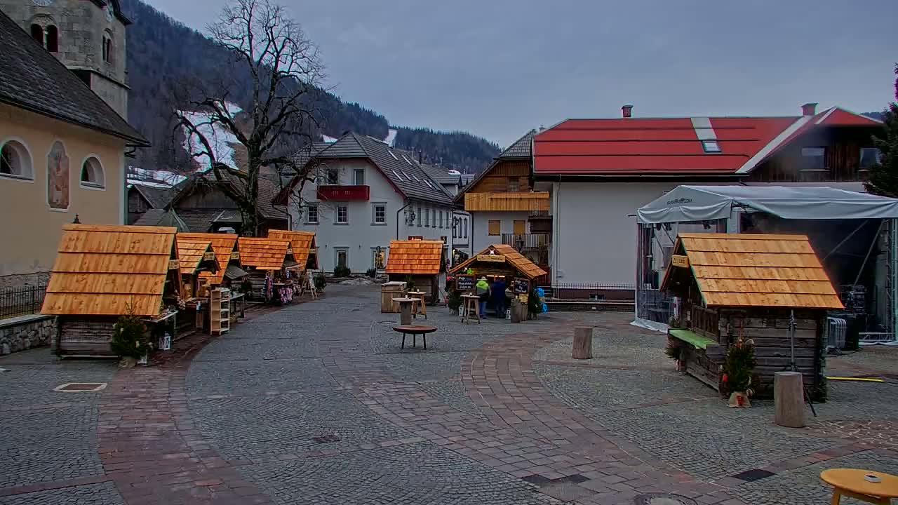 A cobblestone town square with several small wooden market stalls, a few people, and buildings in the background on an overcast day.