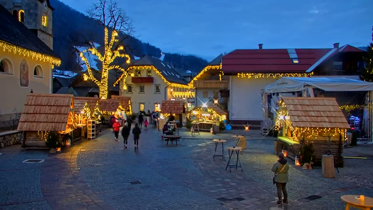 A nighttime view of a European village square with a Christmas market, featuring numerous small wooden stalls illuminated with warm string lights, a large tree also decorated with lights, and people walking around under a twilight sky.