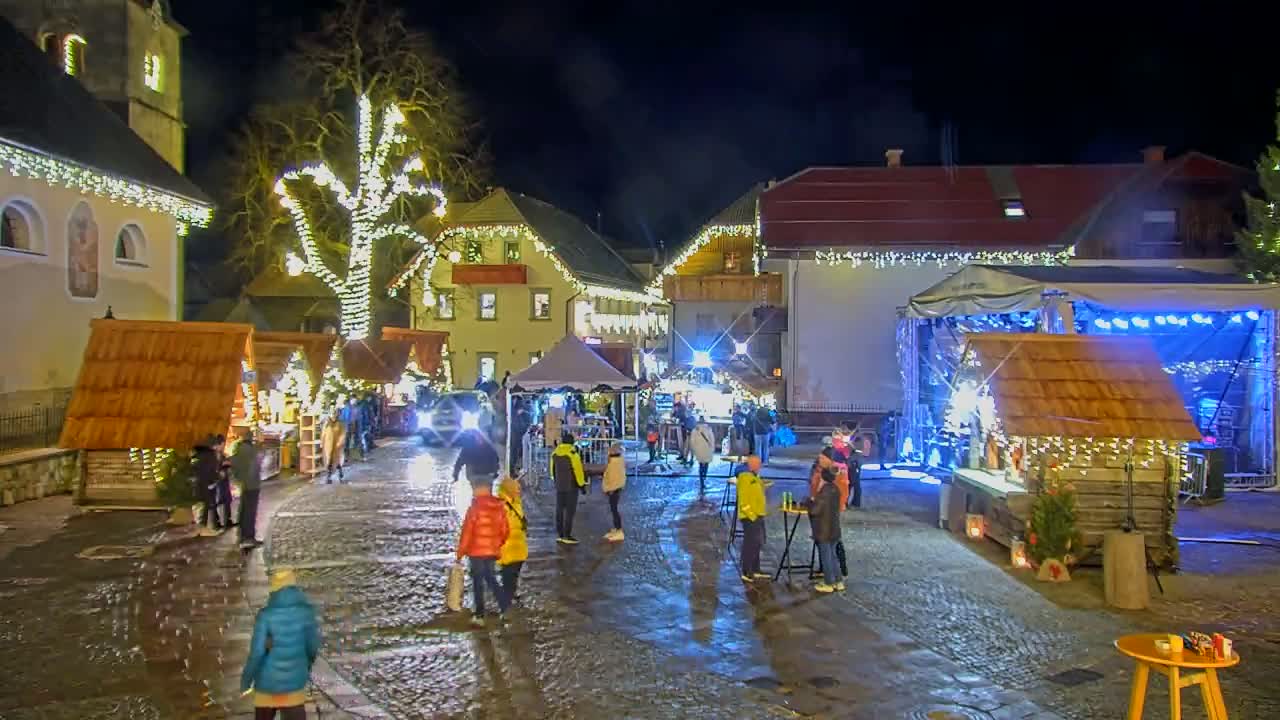 A nighttime outdoor Christmas market scene in a cobblestone town square, featuring wooden stalls with lights, a stage, and people walking around under wet conditions.