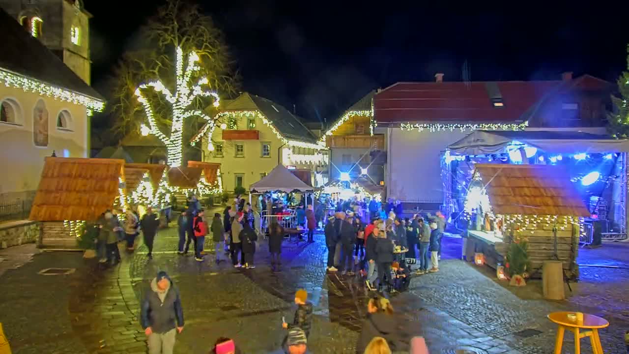 A nighttime outdoor Christmas market scene shows a crowd of people gathered around wooden stalls and a stage with blue lights, illuminated by string lights strung on trees and buildings.