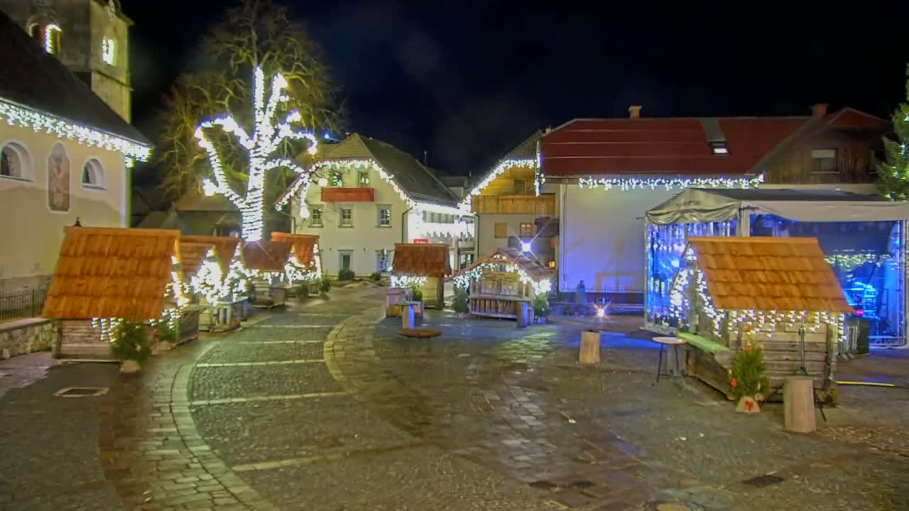 A nighttime scene of a town square adorned with Christmas lights, small wooden huts, and a stage, under a clear, dark sky.