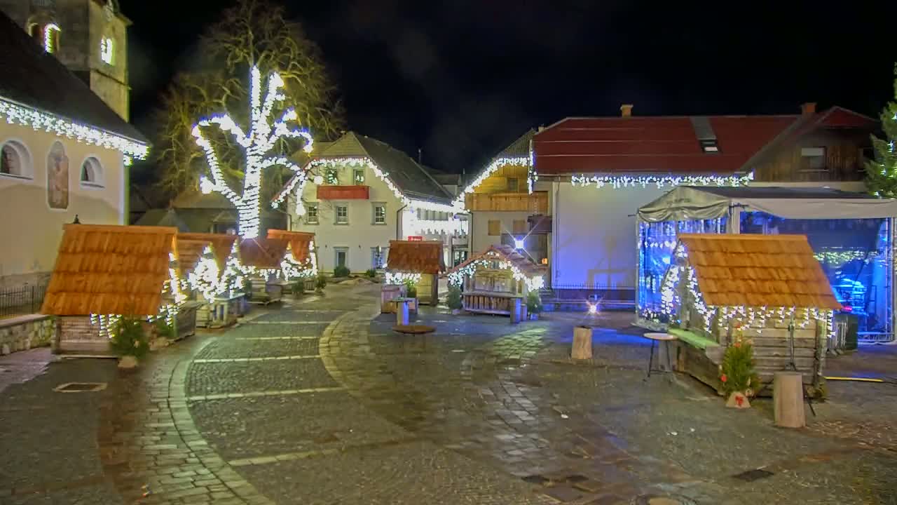 A town square at night is decorated with Christmas lights strung on buildings and small wooden huts, illuminated by the lights and the streetlights.