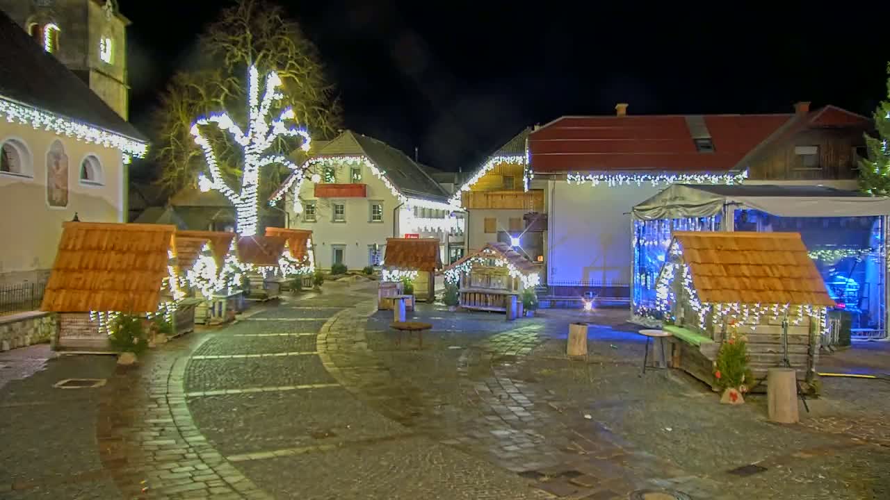 A nighttime view of a town square decorated with Christmas lights, small wooden huts, and a stage.