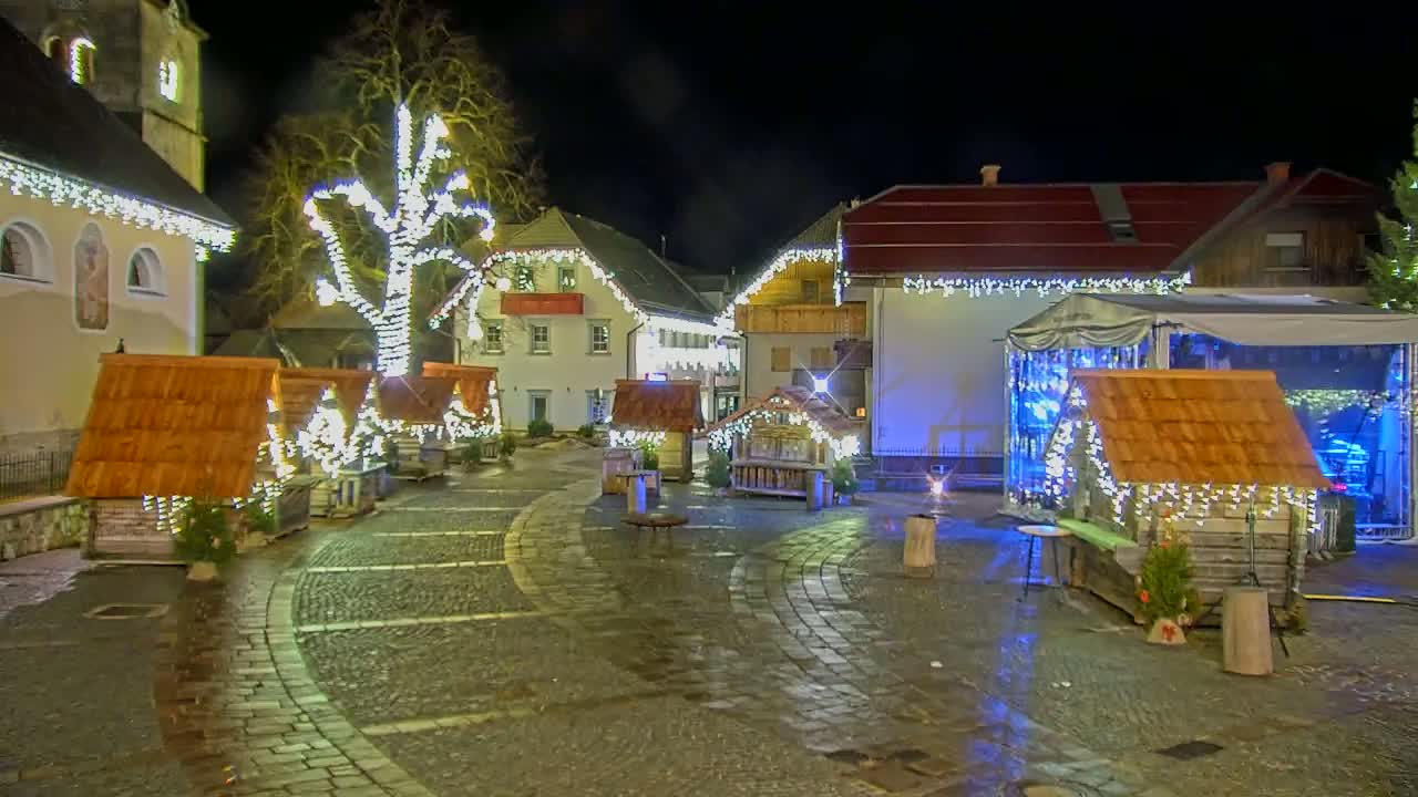 A nighttime view of a town square decorated with Christmas lights, featuring small wooden huts and a large tree illuminated with lights.