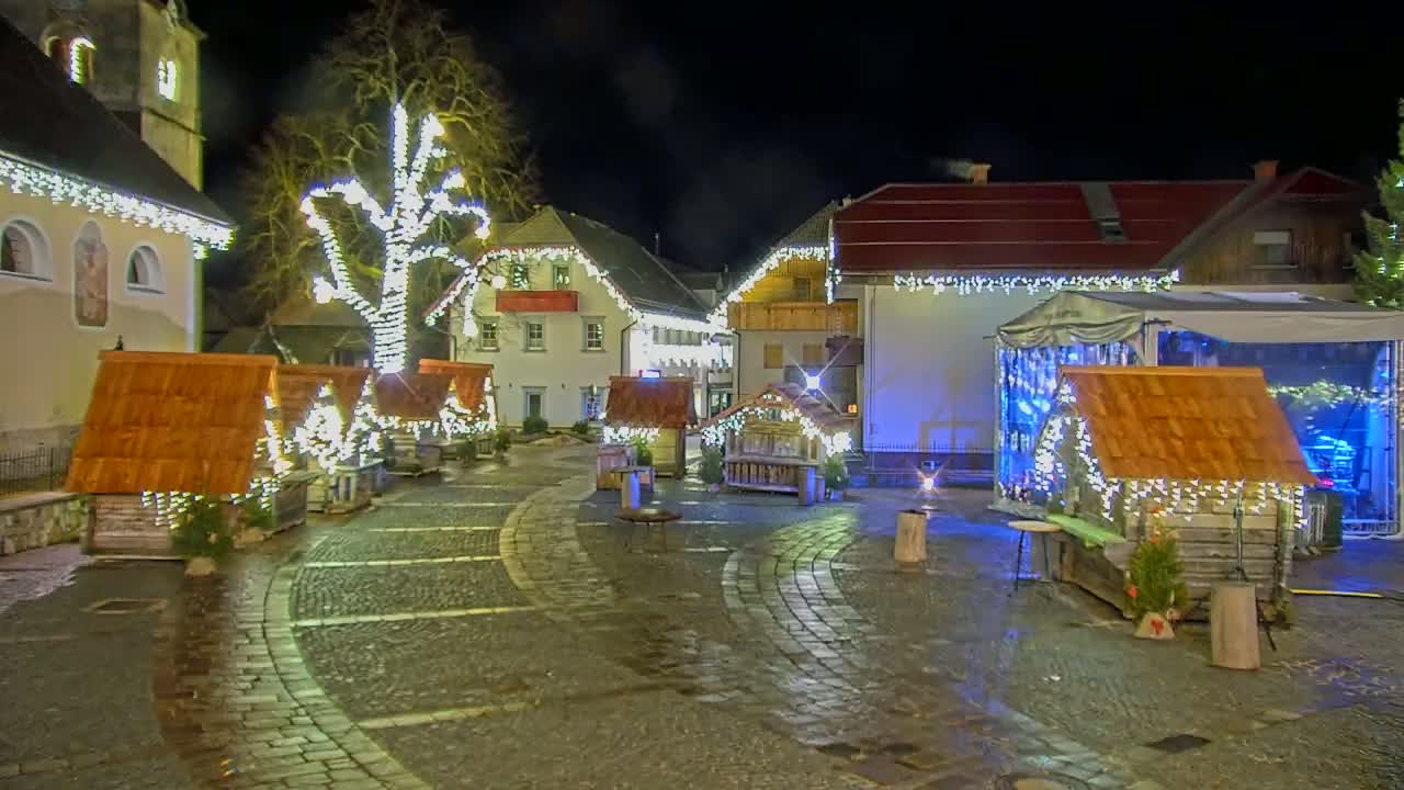 A cobblestone town square at night is decorated with small wooden huts and string lights, illuminated by artificial light.