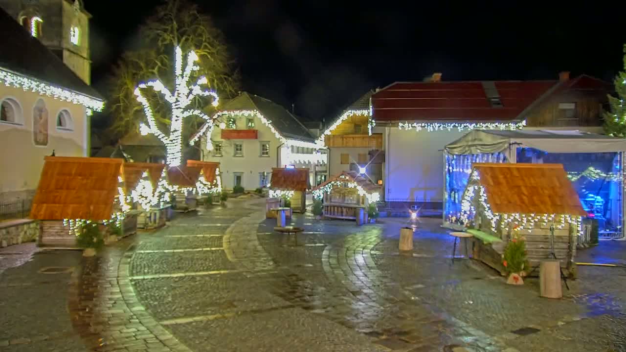 A nighttime scene of a town square decorated with Christmas lights and small wooden huts, with a wet cobblestone surface.