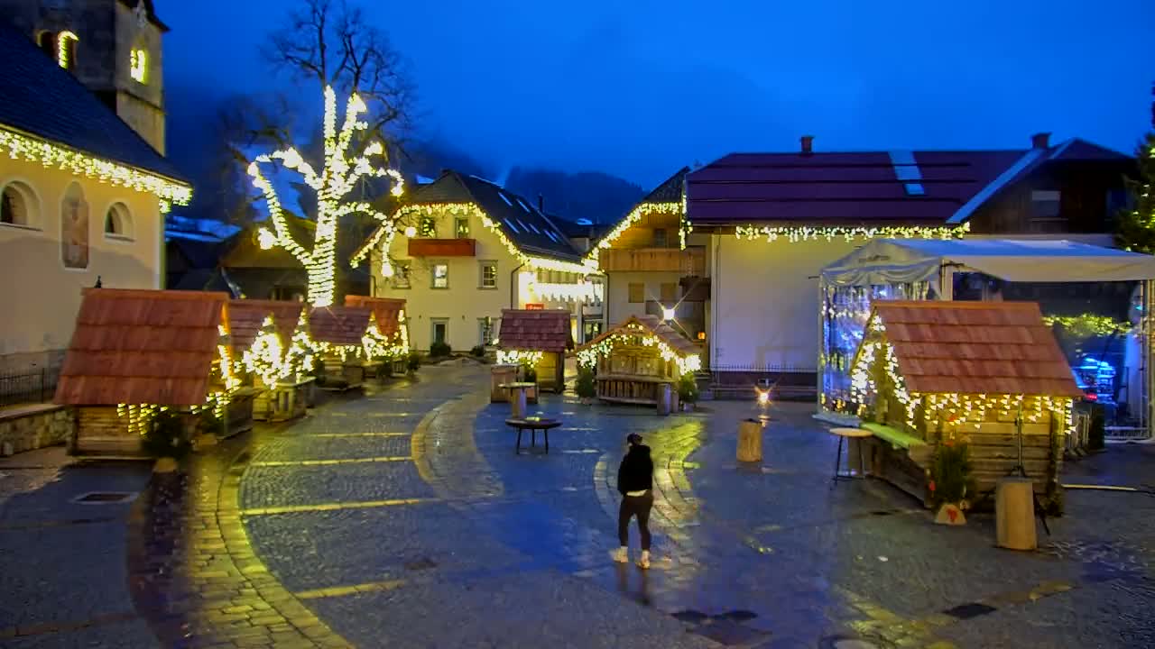 A person stands in a town square at night, surrounded by small, illuminated wooden huts and buildings decorated with string lights, under a dark blue sky.