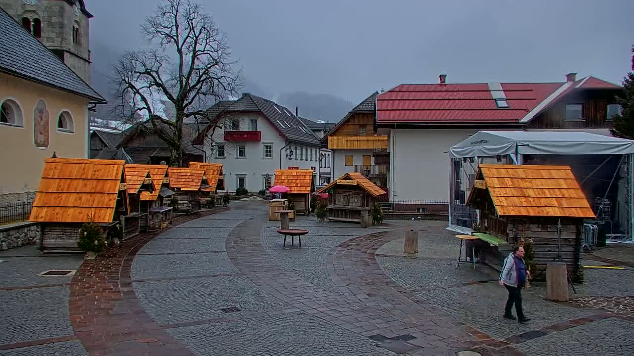 A town square with several small wooden structures, a church, and buildings in the background on an overcast day.