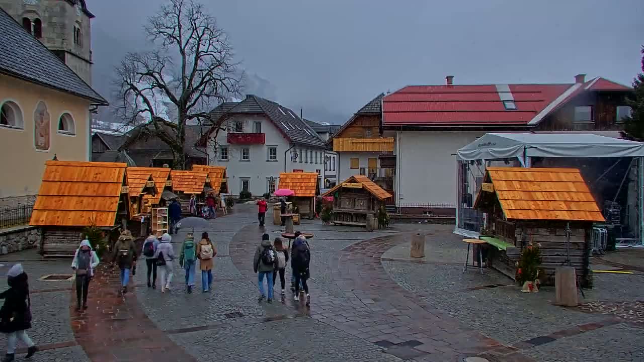 A town square with several small wooden stalls, some people walking around, and buildings in the background under an overcast sky.