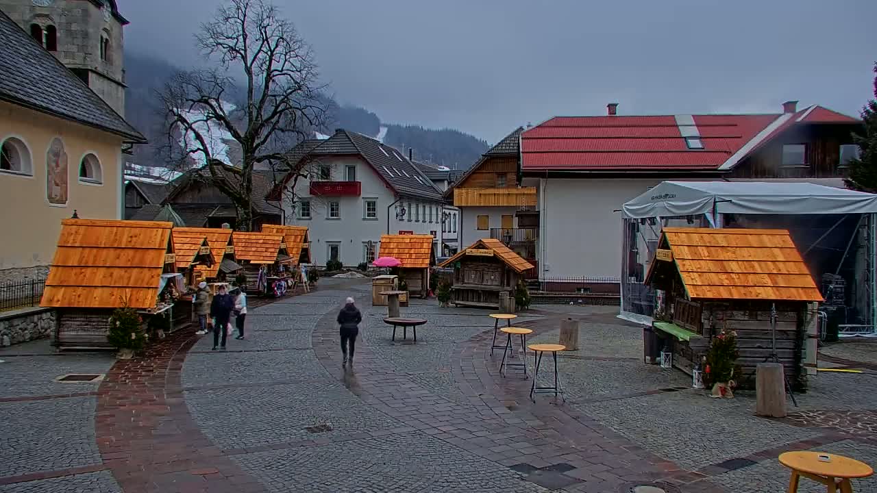 A small, cobblestone town square with several wooden market stalls, a few people walking, and buildings in the background under an overcast sky.