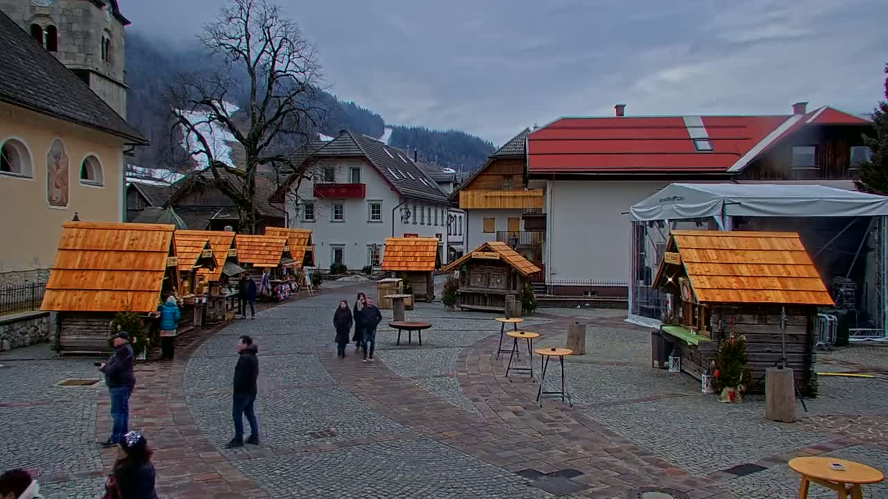 A town square with several small wooden stalls, a few people walking around, and buildings in the background under an overcast sky.