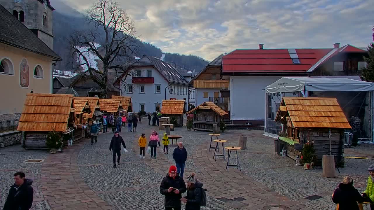 A town square with several small wooden market stalls, people walking around, and buildings in the background under a cloudy sky.