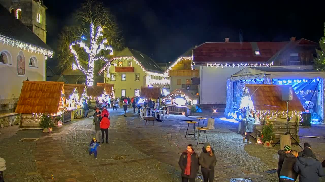 A nighttime outdoor Christmas market is set up in a town square, featuring wooden stalls, a stage, and a large tree decorated with lights.