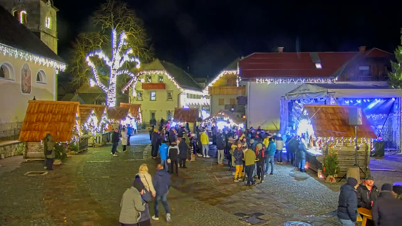A nighttime outdoor Christmas market scene features numerous wooden stalls, a stage, a large crowd of people, and buildings decorated with lights.