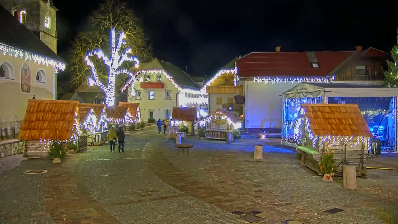A nighttime outdoor scene shows a town square decorated with Christmas lights and small wooden market stalls, with a large tree also adorned with lights in the center.