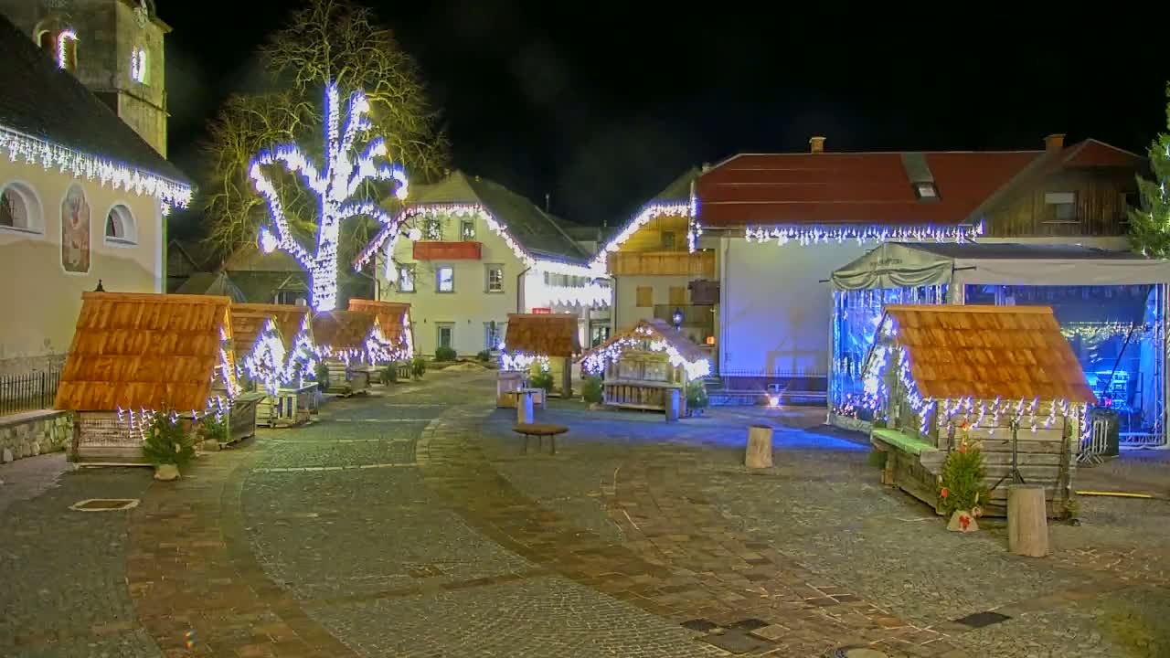 A nighttime town square is decorated with small wooden huts and string lights, illuminated by artificial light.
