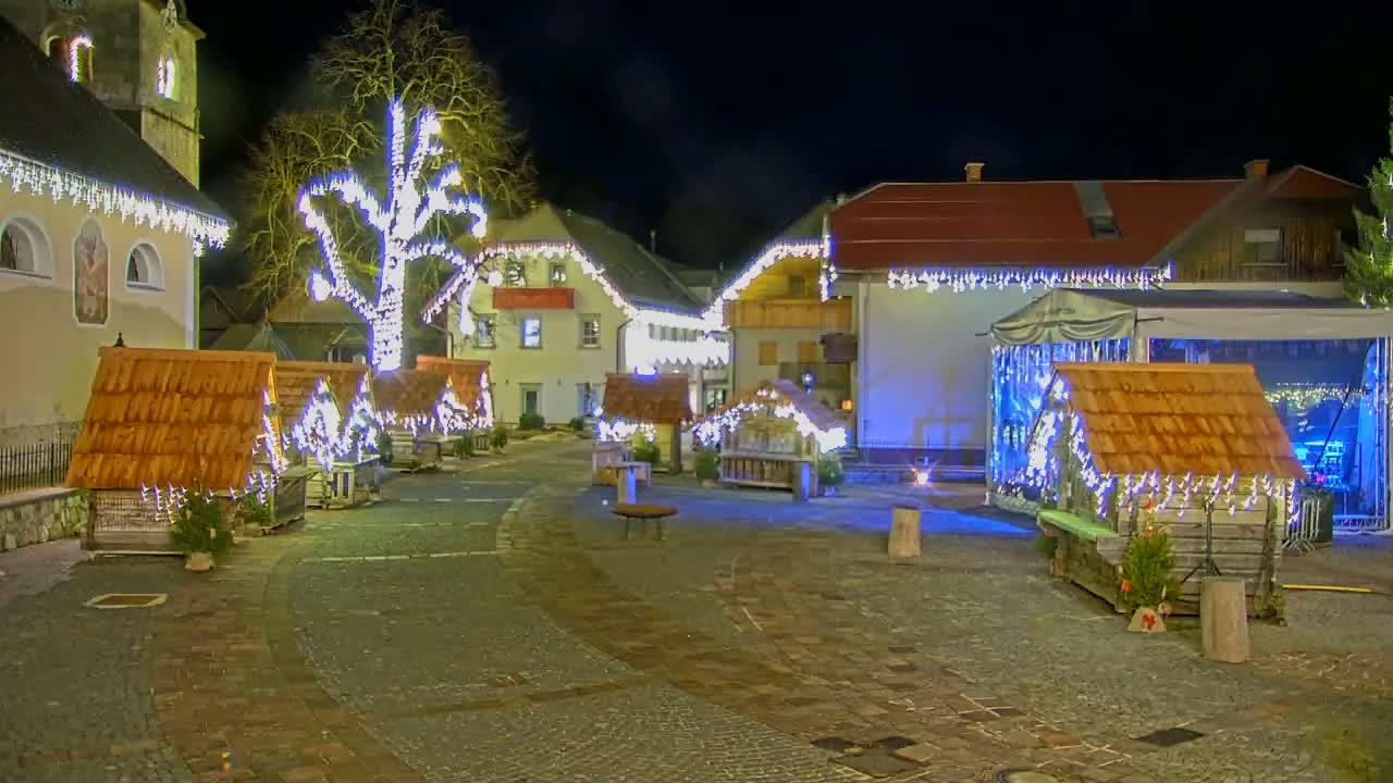 A nighttime scene shows a village square decorated with Christmas lights, small wooden huts, and a large tree also adorned with lights.