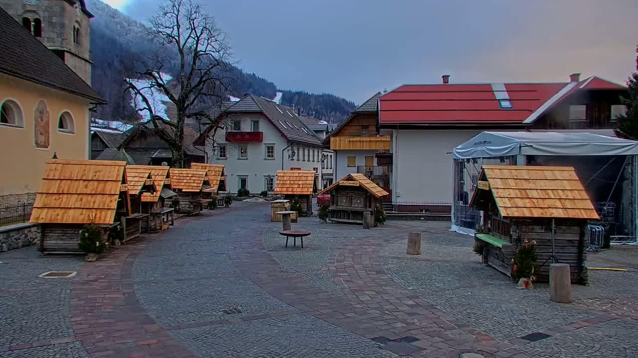 A cobblestone town square features several small wooden stalls with wood-shingle roofs, situated against a backdrop of buildings and snow-covered mountains under an overcast sky.