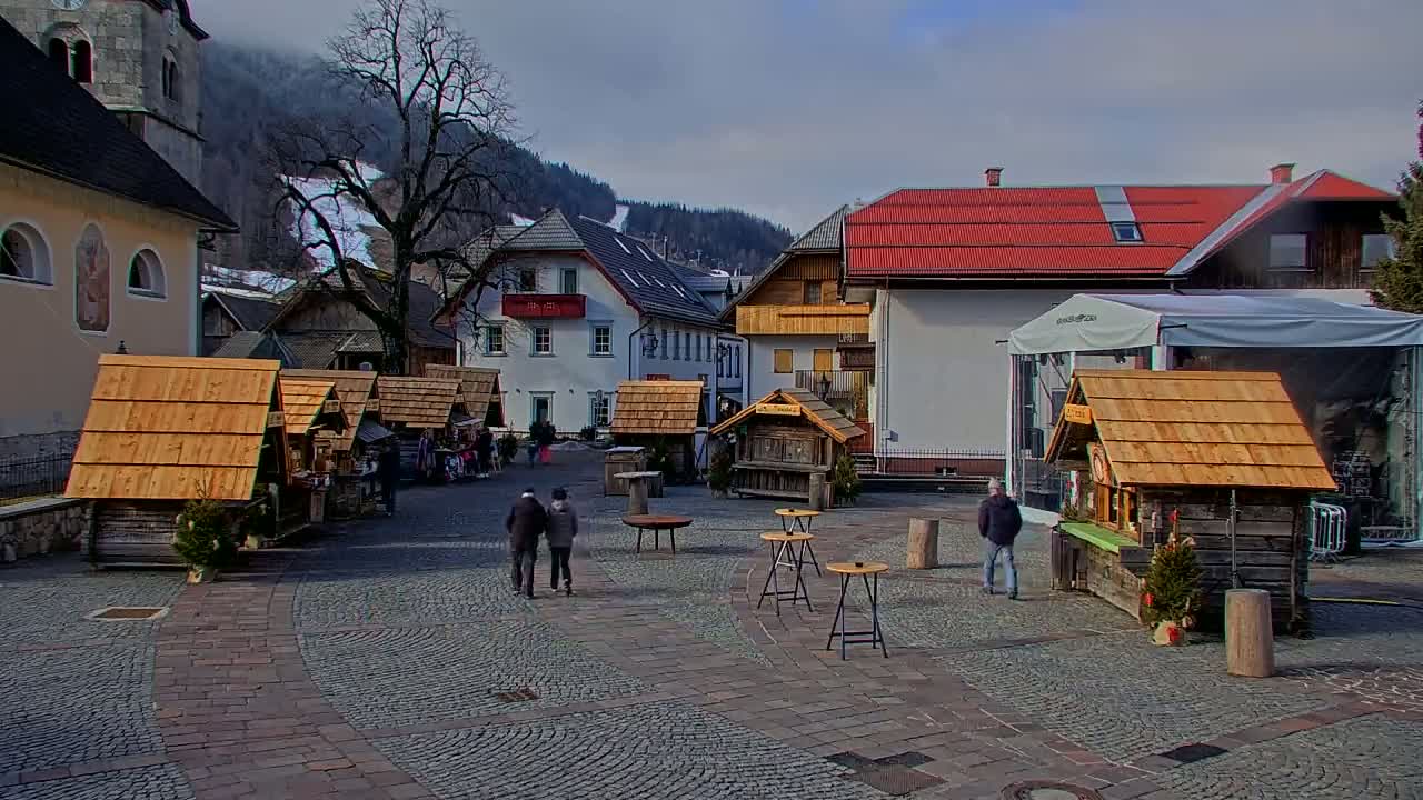 A cobblestone town square features several small wooden stalls, a few people walking around, and buildings in the background, some with red roofs, under a mostly cloudy sky.
