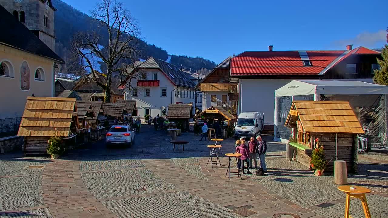 A sunny day in a cobblestone European town square with several wooden market stalls, a few people gathered, and snow-covered mountains in the background.