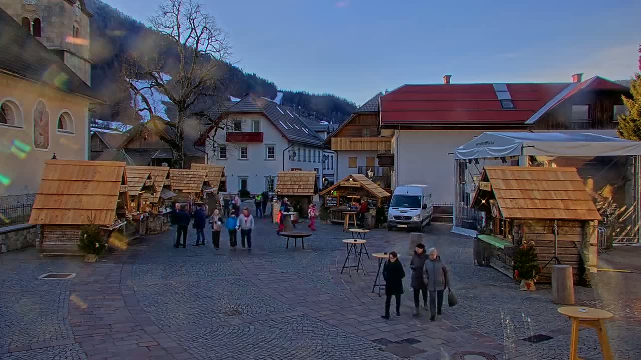 A town square with several wooden market stalls, people walking around, and buildings in the background under a clear evening sky.