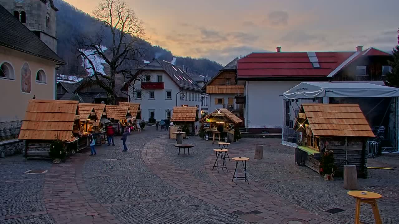 A cobblestone town square at dusk features several small wooden market stalls, a few people, and buildings with a mountain backdrop under a partly cloudy sky.