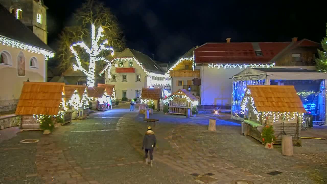 A nighttime scene of a town square decorated with Christmas lights and small wooden huts, with a person walking in the foreground.