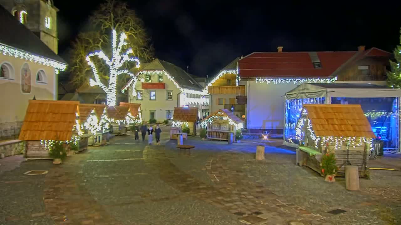 A nighttime scene of a town square decorated with Christmas lights, small wooden huts, and a stage, under a dark sky.