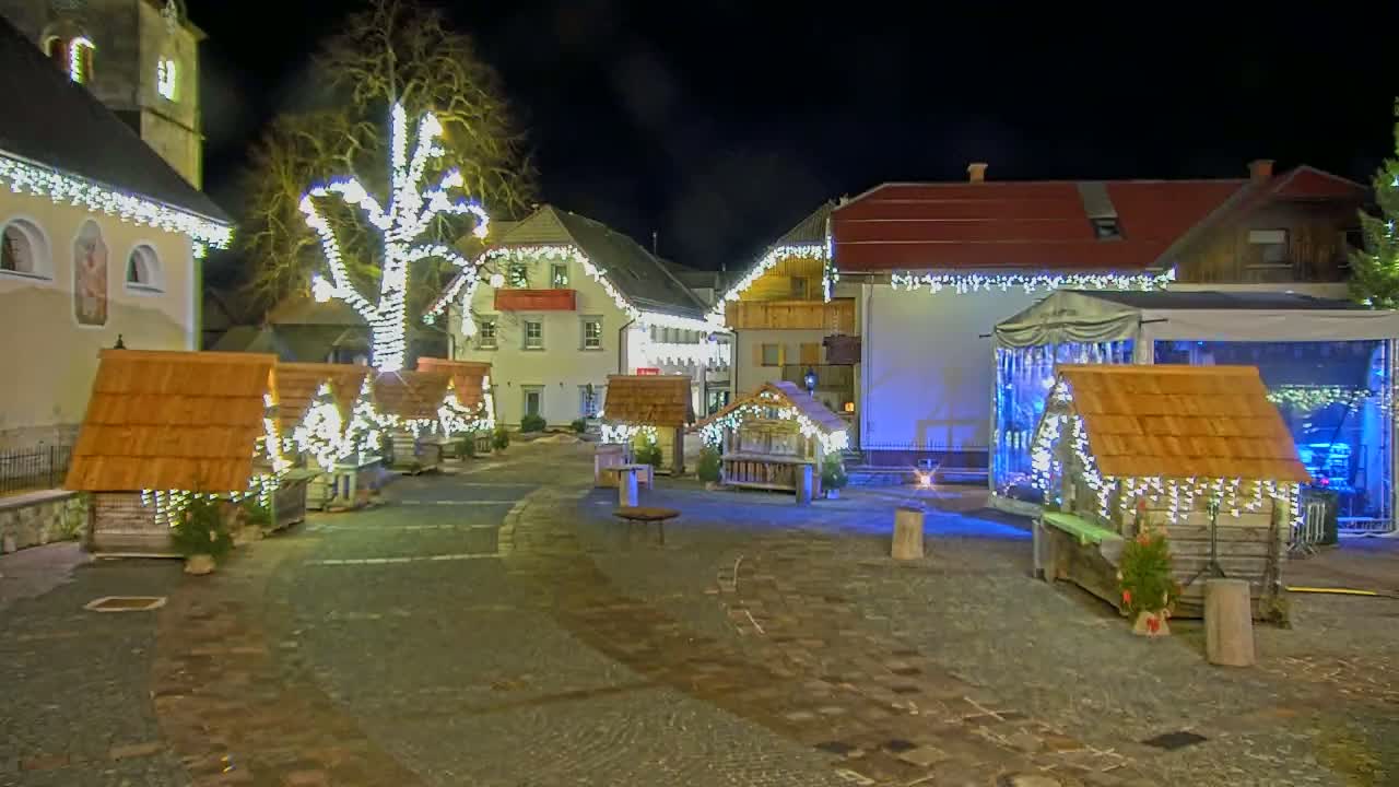A town square at night is decorated with Christmas lights and small wooden structures, some with light displays, near a church and buildings with string lights.