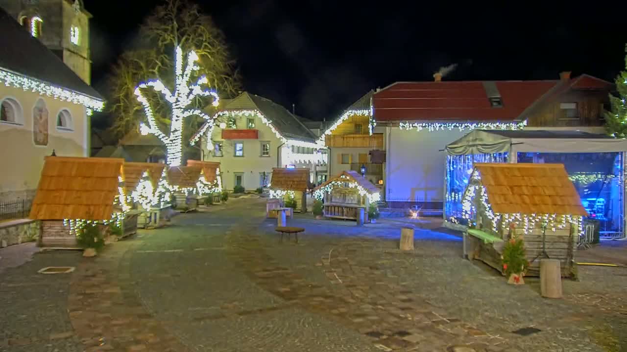 A nighttime village square is decorated with Christmas lights strung on buildings and small wooden huts.