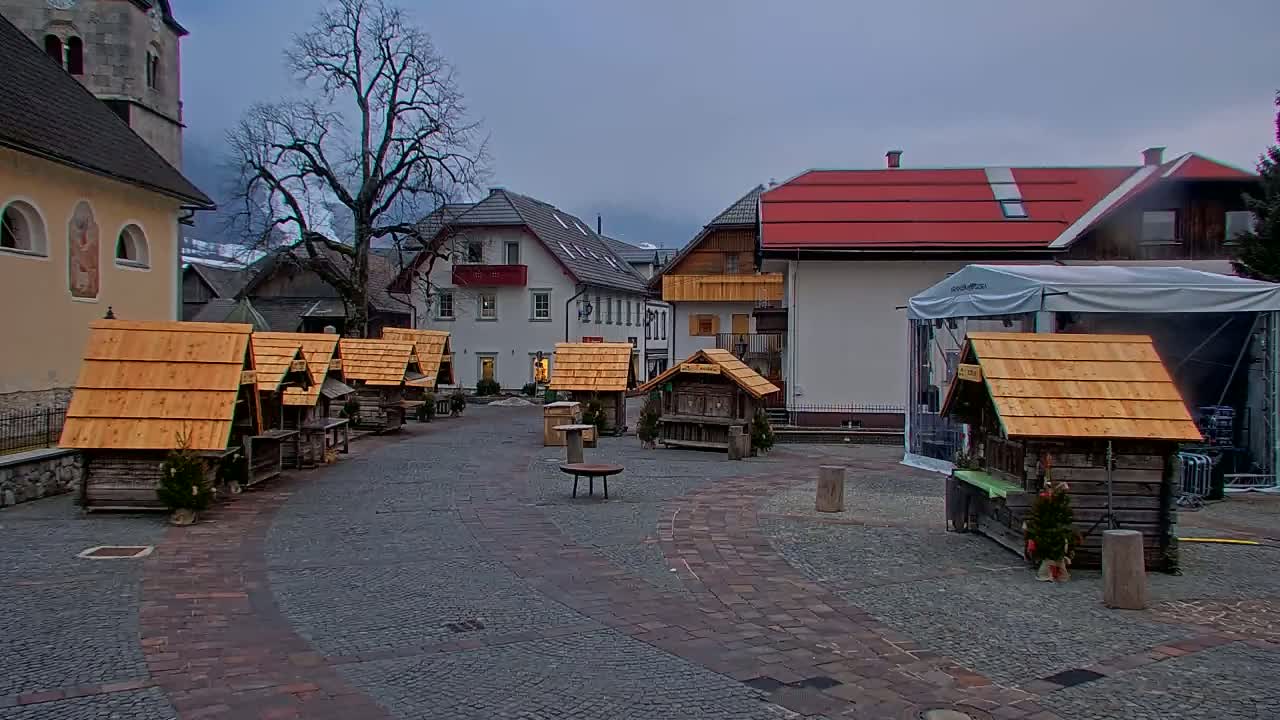 A cobblestone town square under an overcast sky features several small wooden market stalls and buildings, including a church and taller buildings with red roofs.