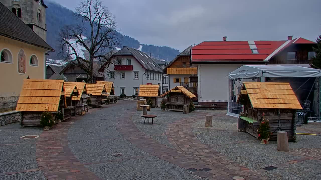 A cobblestone town square, surrounded by buildings and several small wooden stalls with pitched roofs, sits under an overcast sky against a backdrop of snow-dusted mountains.