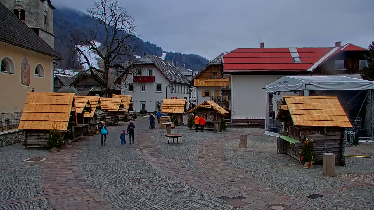 A paved town square with several small wooden market stalls, a few people walking around, and buildings including a church in the background under an overcast sky.