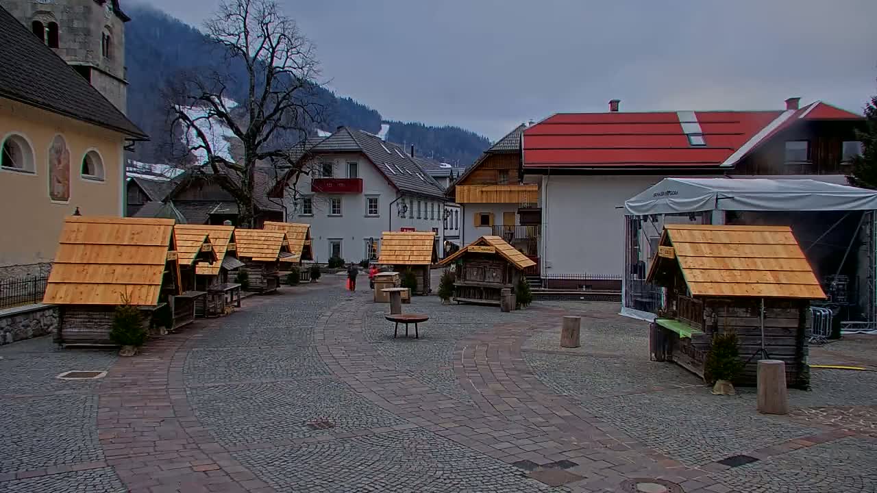 A cobblestone town square with several small wooden stalls, surrounded by buildings and a snow-covered mountain in the background under an overcast sky.
