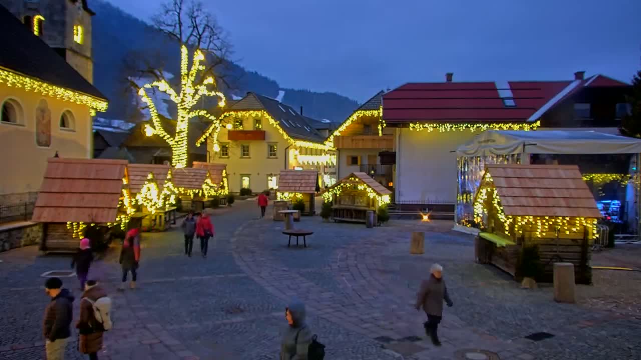 A town square at night is illuminated with string lights adorning buildings and small wooden structures, with people walking around under a twilight sky.