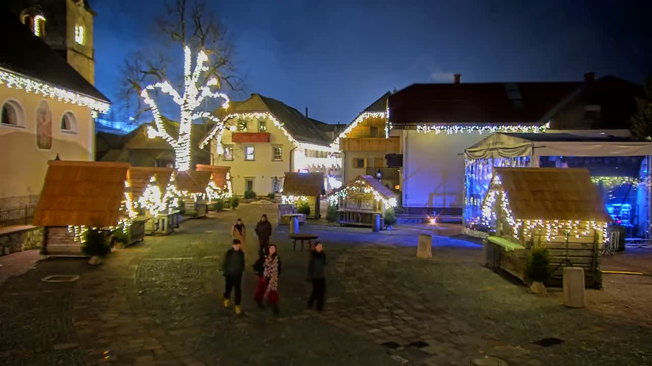 A nighttime scene of a town square decorated with Christmas lights, small wooden huts, and a large tree, with a few people standing in the square.