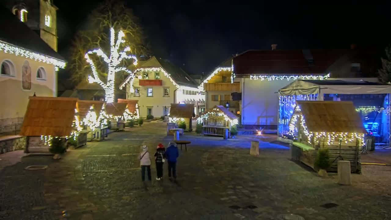 A nighttime view of a town square adorned with Christmas lights and small wooden huts, with three people walking through the square.