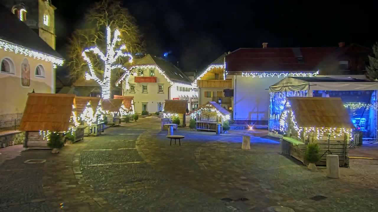 A nighttime scene of a town square decorated with Christmas lights and small wooden huts, a stage, and a large tree.