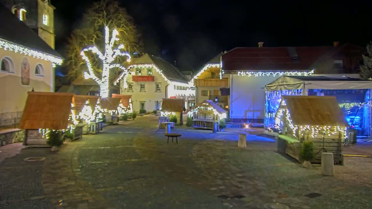 A nighttime scene of a town square decorated with Christmas lights and small wooden huts, with a stage set up in the background.